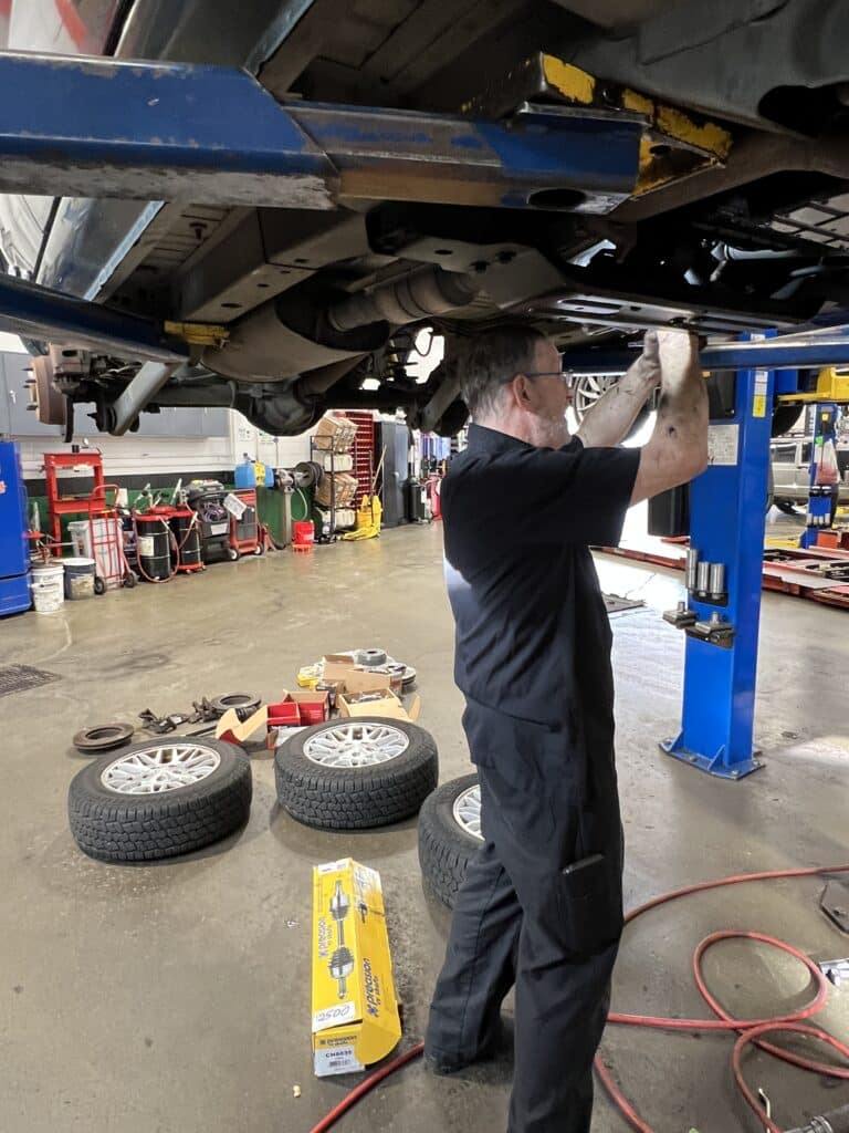 Technician working underneath a Jeep Grand Cherokee during front driveline and steering repairs.